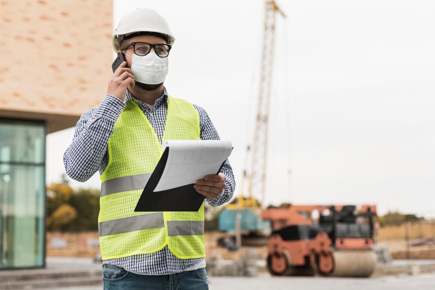 Engineer in hard hat and mask on the phone holding a tablet at a construction site